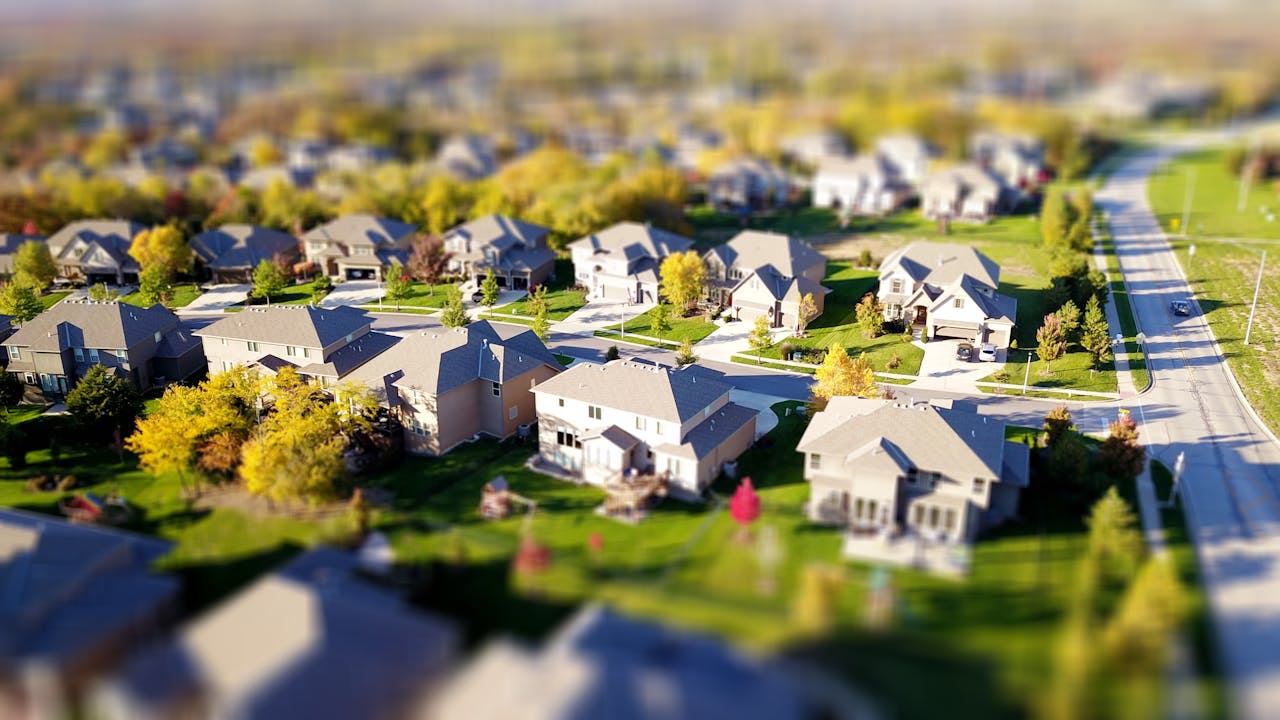 heros-img Tilt-shift aerial photo of a suburban neighborhood with green lawns and roads.